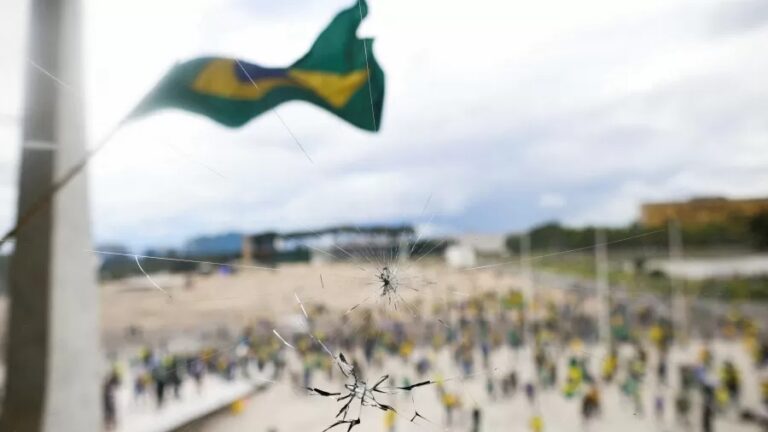 Imagem de vidraça atingida em primeiro plano e em segundo plano manifestantes com efeito desfocado.